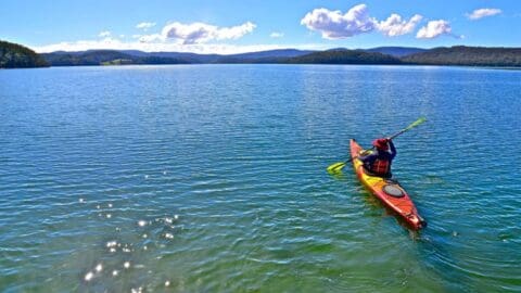 A person kayaks on a pristine lake under a clear blue sky, surrounded by distant hills and calm water.