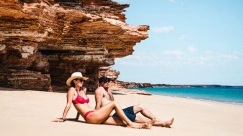 Two people sit on a sandy beach near rock formations and turquoise water, enjoying a sunny day at an eco-resort. The person on the left wears a red bikini and sunhat, while the person on the right wears dark shorts and a cap.