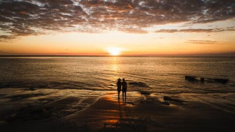 Two people stand on a serene eco-resort beach, facing a sunset over the ocean with clouds scattered across the sky.