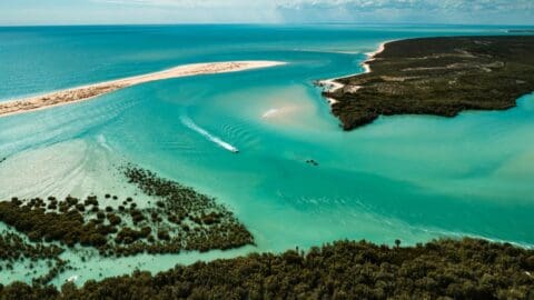 Aerial view of a turquoise ocean inlet with a boat creating a wake. The scene features lush greenery, a sandy shoreline, and an inviting resort nestled along the beach.