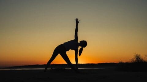 A person practices yoga in a triangle pose against a sunset backdrop on the sands of an eco beach resort.