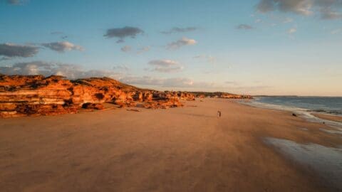 Wide sandy beach with red rocky cliffs on the left, calm ocean on the right, and a person walking in the distance under a clear sky with scattered clouds. Perfect for those seeking a peaceful retreat, this Eco Beach is an ideal resort escape.