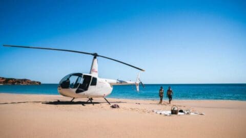 An eco-friendly helicopter is parked on a sandy beach near the ocean, with two people standing nearby and a picnic setup in the foreground.