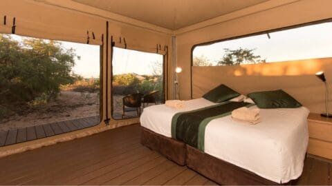 Interior of an eco-glamping tent with a neatly made double bed, green accent pillows, folded towels, bedside lamps, and large windows overlooking a natural, bushy landscape.