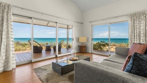 A modern, eco-friendly living room with large glass doors opens to a wooden deck overlooking a scenic ocean view. The room features a gray sofa, a glass coffee table, and deck chairs, creating the perfect beach resort vibe.