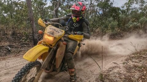 A motorcyclist wearing protective gear rides a yellow dirt bike through a nature-filled, dusty forest trail as part of an adventurous ecotourism tour.