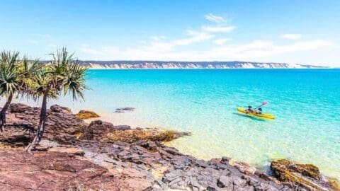 A person paddles a kayak in clear turquoise water near a rocky shore with tropical plants under a bright blue sky, exploring the area as part of an exciting tour.