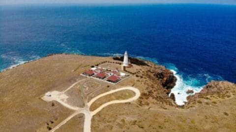 Aerial view of a coastal landscape with a lighthouse, several buildings offering accommodation, winding paths for scenic tours, and the ocean in the background.