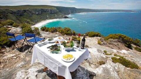 Outdoor dining setup with a table covered with a white cloth, two chairs, and food and wine, positioned on a rocky cliff overlooking a coastline with blue water and cliffs in the background—perfect for those who appreciate nature.