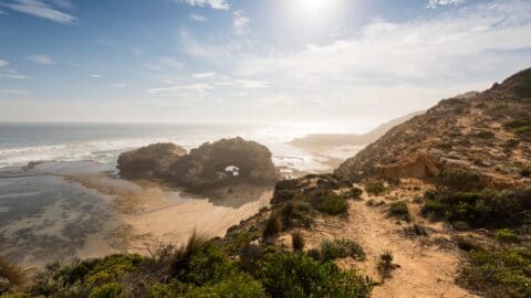 A coastal landscape with rocky formations, sustainable greenery, and a sandy area near the water under a bright sky with scattered clouds, perfect for ecotourism.