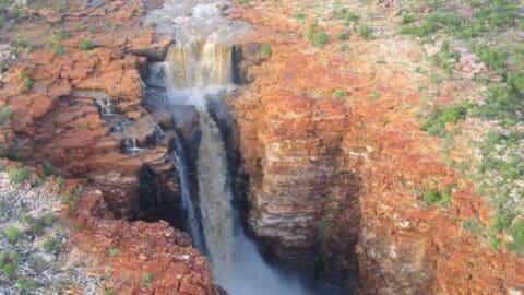 Aerial view of a waterfall flowing through a deep, rocky gorge surrounded by reddish-brown cliffs and lush vegetation, perfect for an adventurous tour.