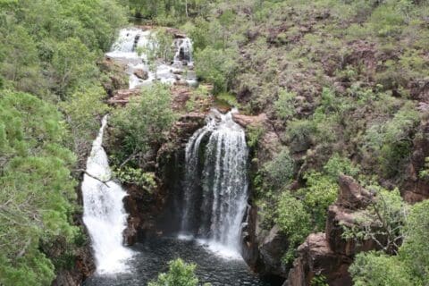 A multi-tiered waterfall cascades over rocky cliffs surrounded by lush green vegetation and trees, offering an enchanting glimpse into the Northern Territory's natural beauty. Enhance your experience with Indigenous Tours that reveal the rich cultural history of the land.