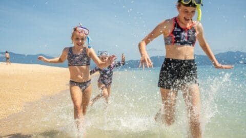 Three children in swimsuits and snorkeling gear play and splash in the shallow water at a sandy beach with a backdrop of mountains, enjoying their time near an ecotourism accommodation.