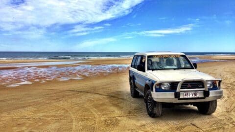 A white Mitsubishi SUV drives on a sandy beach with the ocean waves and blue sky in the background, offering a sustainable tour experience.