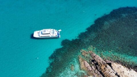 Aerial view of a white boat anchored in clear turquoise water near a rocky shoreline, offering an ideal spot for a tour of the area.