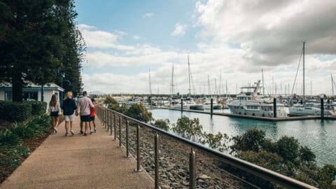 People walk along a paved path beside a marina with numerous boats docked. The sky is partly cloudy, and there are trees and shrubbery along the path.