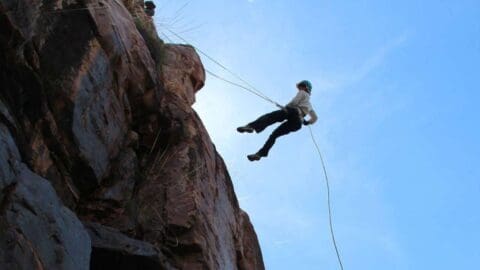 A certified person is mid-air, descending by rappelling down a rocky cliff, wearing a helmet and safety gear against a clear blue sky.