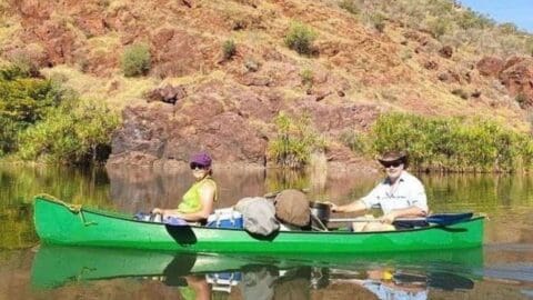 A woman and a man paddle a green canoe on a calm body of water, surrounded by rocky and vegetated terrain. The canoe, loaded with supplies, reflects their commitment to sustainable adventure.