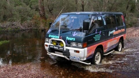 A van with "Grampians Tours" written on it drives through a shallow creek in a forested area, offering an immersive nature tour experience.