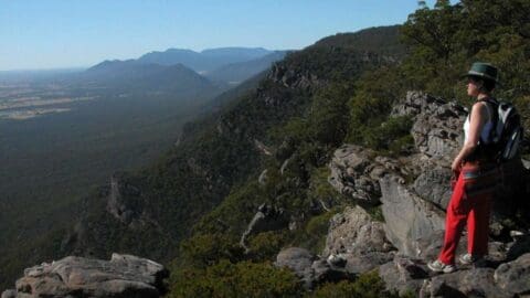 A person in red pants and a hat stands on a rocky outcrop overlooking a vast, forested mountain range, offering an ideal spot for ecotourism enthusiasts seeking sustainable adventures.