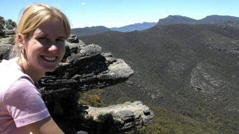 A person with blonde hair smiles while sitting on a rocky ledge overlooking a vast mountainous landscape under a clear sky, enjoying an ecotourism tour.