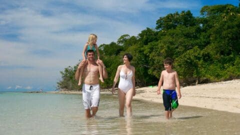 A family of four, two adults and two children, enjoy a sunny day walking along the shoreline of a beach. The man carries a young girl on his shoulders while the woman and an older boy walk beside them, exploring the coastline on their sustainable tour.