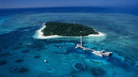 Aerial view of an island with lush greenery, surrounded by clear blue waters and coral reefs. A pier extends from the island with boats docked nearby, offering sustainable accommodations for eco-friendly travelers.