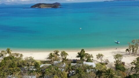 Aerial view of a coastal area with clear blue water, sandy beach, scattered trees, small boats, and distant islands. Several buildings and structures are visible near the shoreline. Perfect for an ecotourism tour that highlights sustainable practices.