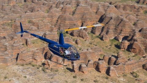 A blue helicopter flies over a landscape of layered rock formations with distinct stripes, showcasing the beauty of ecotourism.