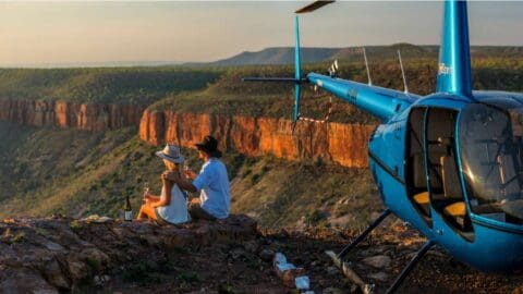 A couple sits by a blue helicopter on a rocky ledge overlooking a vast canyon landscape during sunset, experiencing sustainable ecotourism.