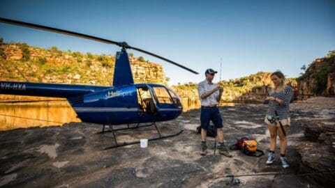 Two people stand near a blue "HeliSpirit" helicopter by a rocky riverbank, preparing fishing gear. Rugged cliffs and calm water are in the background, showcasing the beauty of ecotourism in nature.