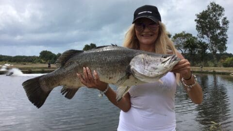 A person wearing sunglasses and a hat holds a large fish by a lakeside, with trees and cloudy sky in the background, enjoying the serene nature.