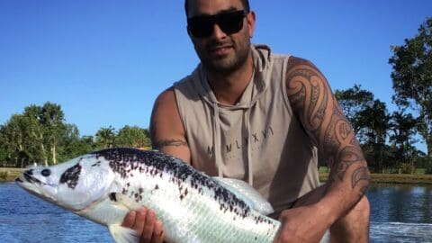 A man with tattoos and sunglasses holds a large fish in front of a body of water and lush trees under a clear blue sky, offering an authentic tour experience.