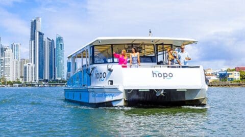 A Hopo ferry sails on a waterway with several people on board, and tall city buildings are visible in the background, creating a picturesque scene for The Tour Collective.