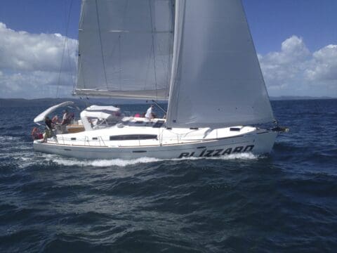 A large white sailboat named Blizzard glides on a sunny day in the open waters of the Whitsundays, with several people onboard, enjoying their travel adventure.