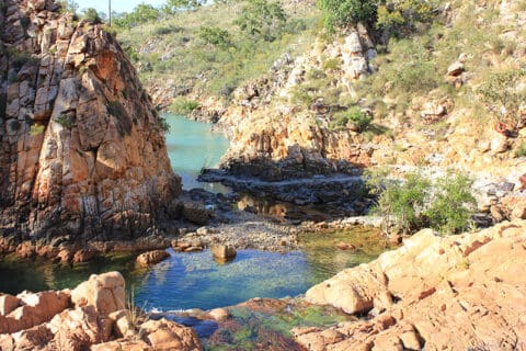 A narrow river flows through a rocky, arid landscape with sparse vegetation and steep, jagged cliffs on either side, reminiscent of the rugged beauty found beyond the bustling waters of Sydney Harbour.