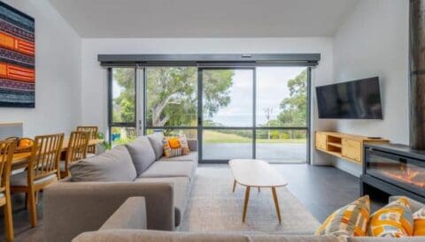 Modern living room with a gray sofa, wooden coffee table, wall-mounted TV, dining table with chairs, large windows showcasing an outdoor scene in Lorne’s Bush House Cottages, and a fireplace. Bright decor accents the space.