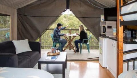 Two people sit at a small table on a balcony in a glamping tent at Bush House Cottages, with a view of trees in the background. They are engaged in conversation. The interior includes a couch, bed, and kitchenette, making this Eco Retreats destination near Lorne perfect for relaxation.