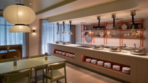 Modern dining area at Ibis Styles in central Sydney, featuring tables, chairs, and a well-lit buffet counter with assorted cookware and dishes. Decor includes hanging lamps, large windows, and open shelving with orange accents.