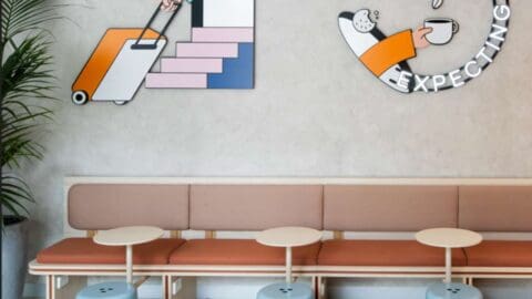 A cozy waiting area at Ibis Styles Central features light brown seating, small round tables, and colorful wall art depicting a suitcase and a cup of coffee. A plant sits on the left side, adding a touch of greenery to this stylish Sydney locale.
