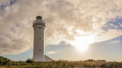 A white lighthouse stands tall in a grassy area under a partly cloudy sky with the sun shining brightly, offering an idyllic spot for ecotourism and sustainable tours.