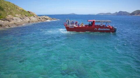 A group of people ride a red sightseeing boat named "Envision" in a clear blue sea with rocky hills and distant islands in the background, embracing the principles of ecotourism.