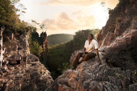 A person sits on a rocky cliffside in the Northern Territory during sunset, while two others walk along a path in the background surrounded by lush vegetation and rocky formations, perhaps exploring on one of the many Indigenous Tours available.