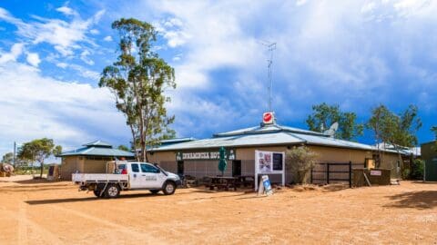A rural building with signage reading 'Birdsville Hotel' under a partly cloudy sky. A white utility vehicle is parked in front of the building, which is surrounded by trees and dirt ground, perfect for nature enthusiasts or an ecotourism tour.