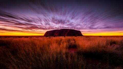 A large rock formation stands in the distance against a vibrant, colorful sunset sky, inspiring thoughts of grand journeys and endless adventures, surrounded by grassland.