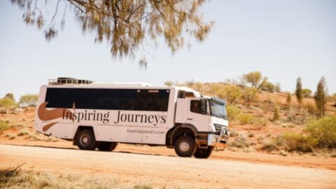 A white bus with "Inspiring Journeys" branding is parked on a dusty road in an inspiring desert landscape with sparse vegetation.