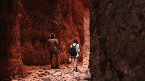 Two people with backpacks embark on inspiring journeys through a narrow, rocky canyon with tall red-brown walls.