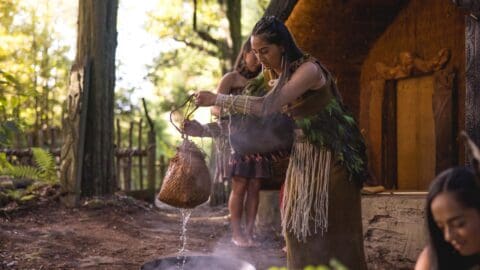 A woman in traditional attire pours liquid from a woven container into a pot outdoors, set against the backdrop of a wooden structure and forest. The scene, filled with other people, captures the inspiring journey of communal practices and cultural heritage.