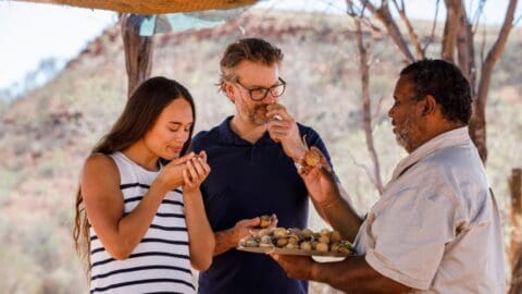 Three people stand outdoors, sampling food from a platter held by a man. They are under an inspiring shade structure with a rocky landscape in the background, evoking the sense of shared journeys and culinary adventures.