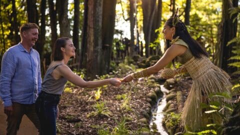 A woman in traditional attire, embodying the spirit of inspiring journeys, exchanges an object with another woman and a man, standing in a lush forest setting.
